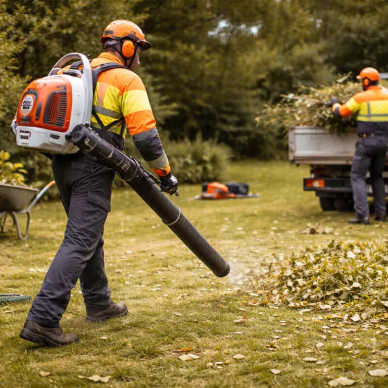 A worker in high-visibility gear clearing leaves on a lawn while cut branches are loaded into a truck